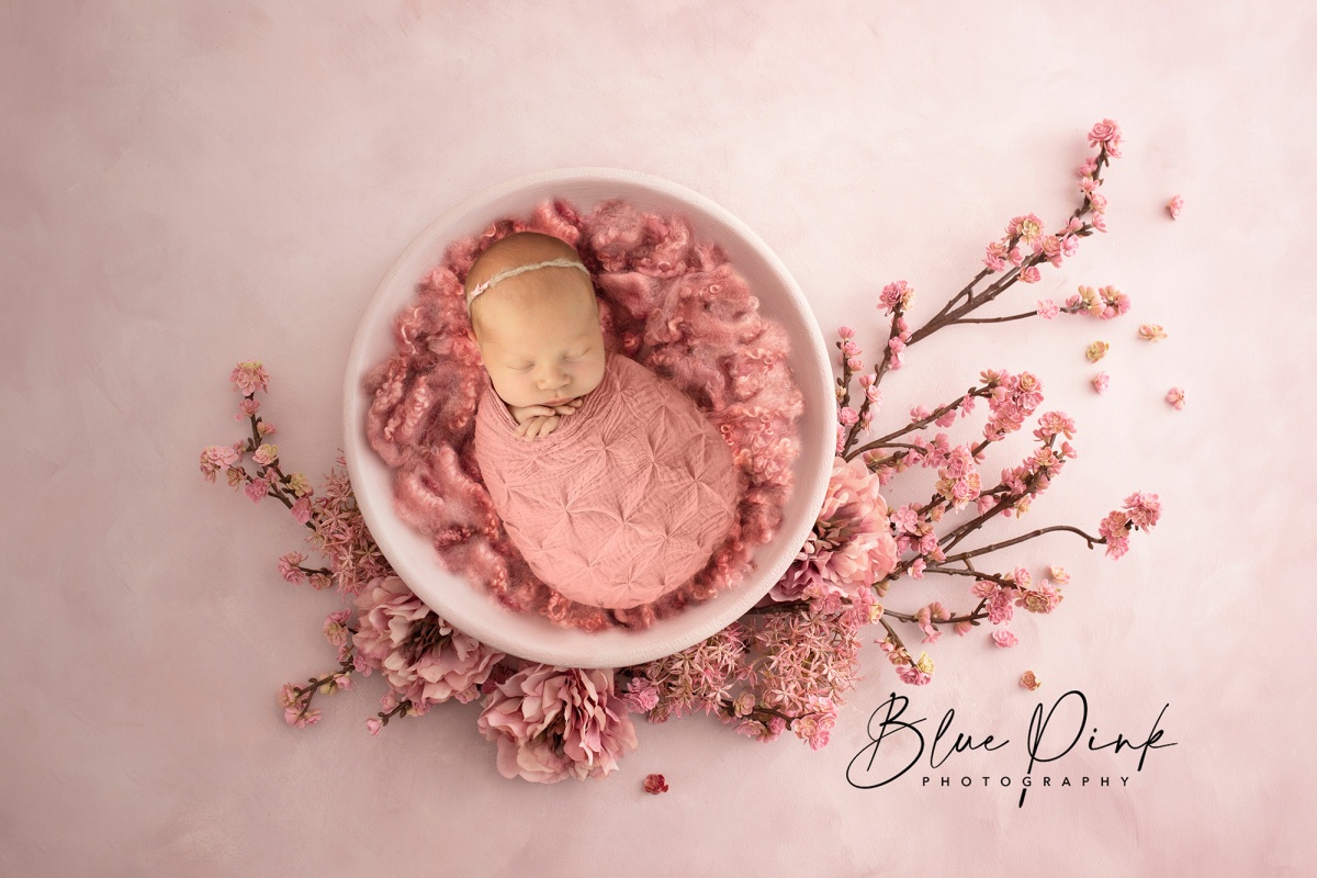 Adorable newborn baby girl, swaddled in soft fabric, nestled in a pale pink bowl adorned with delicate pink flowers, capturing a serene and charming Japanese-inspired scene.